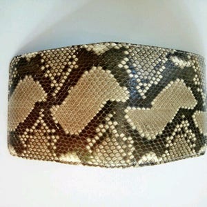 May include: A bi-fold wallet with a snakeskin pattern. The wallet features a repeating pattern of tan, brown, and gray scales. The wallet is rectangular and appears to be made of leather.
