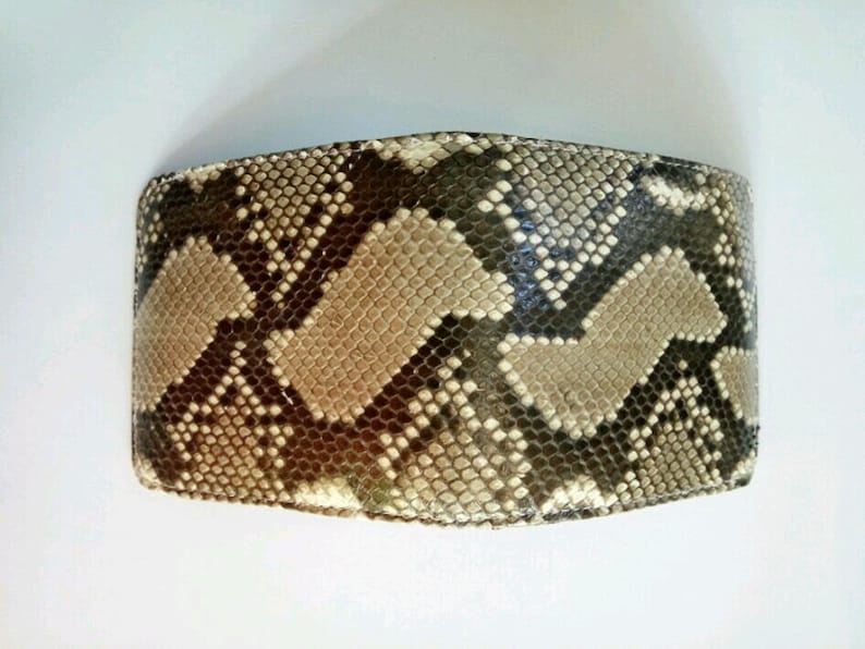 May include: A bi-fold wallet with a snakeskin pattern. The wallet features a repeating pattern of tan, brown, and gray scales. The wallet is rectangular and appears to be made of leather.