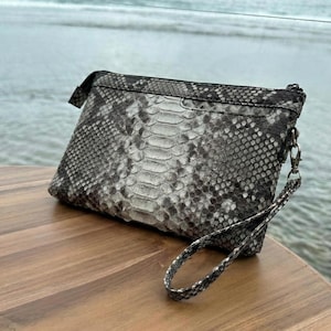 Könnte beinhalten: Eine rechteckige Clutch mit Schlangenmuster in Grau- und Schwarztönen. Sie hat eine Handschlaufe, die mit einem silberfarbenen Verschluss befestigt ist. Der Hintergrund zeigt eine Holzoberfläche mit dem Meer in der Ferne.