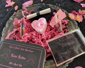 May include: A mini spell kit on a silver tray, featuring a heart-shaped candle, a small vial, and a tin with the text "Love Letter". The kit includes a scroll, a small bottle with dried flowers, and pink paper filler.