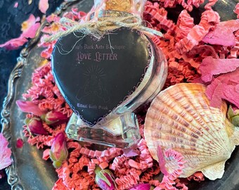 Peut inclure: Une bouteille en verre en forme de cœur avec l'inscription "Love Letter", un bouchon en liège et de la ficelle. La bouteille est posée sur un plateau argenté avec du papier rose, des boutons de rose et un coquillage. La bouteille est remplie d'une substance blanche.