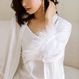 May include: White satin robe with feather trim on the sleeves. The robe has a V-neck and long sleeves.