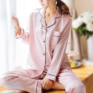May include: A pink satin pajama set with long sleeves and a button-up top. The top has a collar and a pocket on the left side. The pants have a black trim.