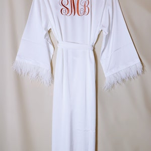 May include: White satin robe with feather trim on the sleeves. The robe has a sash and features a monogram embroidered on the back that says "SMB".
