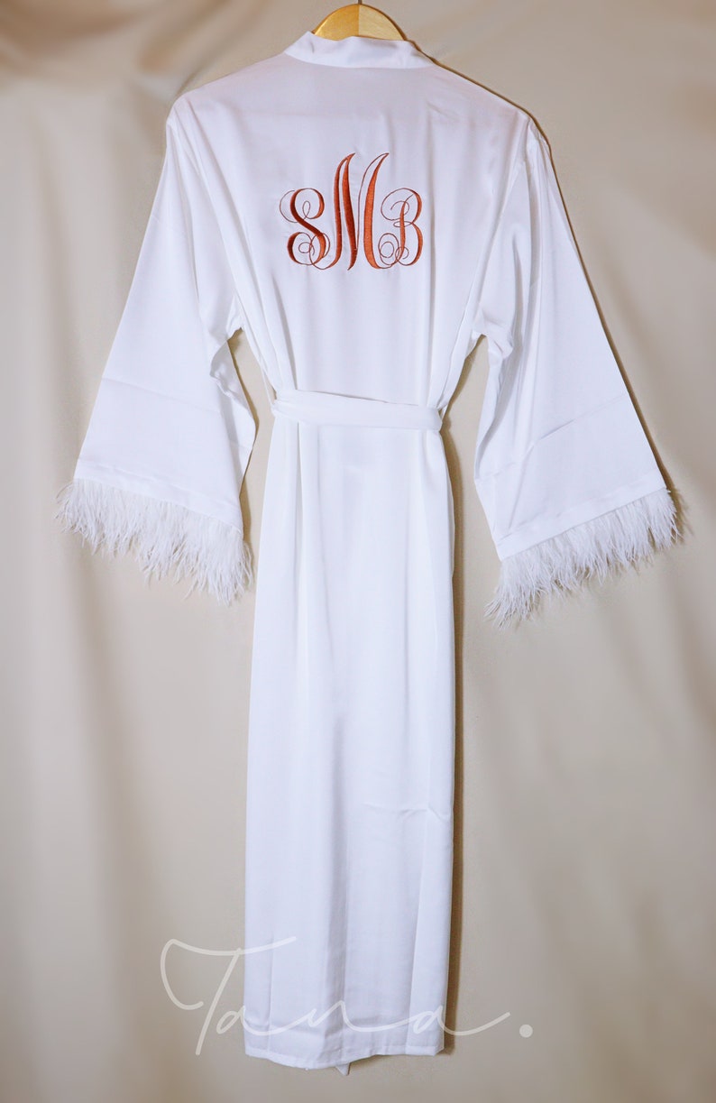 May include: White satin robe with feather trim on the sleeves. The robe has a sash and features a monogram embroidered on the back that says "SMB".