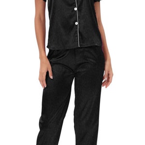 Pode incluir: Conjunto de pijama de cetim preto com mangas curtas e uma parte superior com botões. A parte superior tem uma bainha branca e um bolso do lado esquerdo. As calças têm uma bainha branca na parte inferior.
