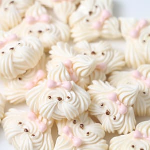 Puede incluir: Una colección de galletas de merengue blancas con forma de perro con lazos rosas y detalles dorados.