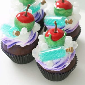 May include: Close-up of several cupcakes with dark brown bases and purple and white frosting. Each cupcake is topped with a red apple, green accents, a small turquoise bottle, and white bubble-shaped decorations. The cupcakes are arranged on a white surface.