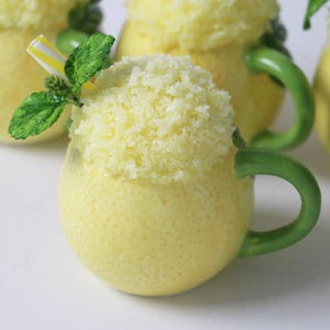 Könnte beinhalten: Vier gelbe Keramikbecher mit grünen Griffen, gefüllt mit einem gelben, fluffigen, geschlagenen Topping. Ein Becher hat ein grünes Blatt und einen gestreiften Strohhalm, der aus dem Topping herausragt.