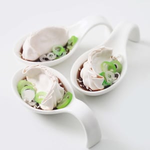 May include: Three white ceramic spoons, each filled with a dumpling, green onion slices, and a dark sauce. The spoons have a curved handle and are arranged on a white surface.