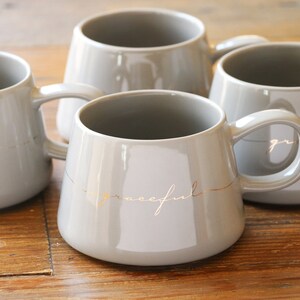 May include: Four gray ceramic mugs with gold lettering. The mugs have rounded handles and the words "soul" and "graceful" are written in gold on the mugs.