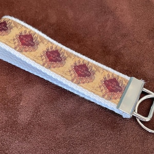 May include: A white and beige key chain with a red and gold patterned ribbon. The ribbon has a repeating design of a red heart with gold accents. The key chain has a silver metal ring.