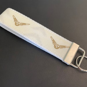 May include: White fabric keychain with a silver key ring. The keychain has two gold-colored illustrations of a winged ball.