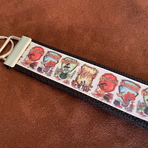 May include: Black keychain with a white ribbon featuring colorful graphics and text. The ribbon displays the names of four houses: Gryffindor, Ravenclaw, Slytherin, and Hufflepuff. A silver-colored metal ring is attached.