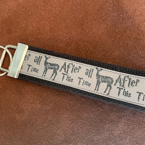 May include: Black and white key chain with a silver ring. The key chain has a black fabric strap with a white ribbon that says "After all this time" with a stag silhouette.