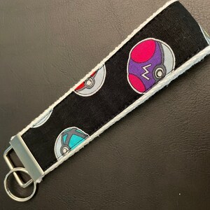 May include: A black and white key chain with a white border. The key chain has a repeating pattern of Poke Balls with different colors. The key chain has a silver key ring.