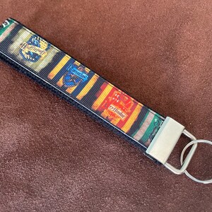 May include: A black and white key chain with a colorful ribbon featuring the Hogwarts house crests: Gryffindor, Hufflepuff, Slytherin, and Ravenclaw. The ribbon is attached to a silver key ring.