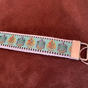 May include: A green, white, and black key chain with a silver key ring. The key chain features a repeating pattern of the four Hogwarts house crests.