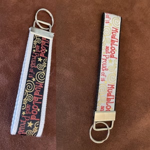 May include: Two keychains with black and white ribbon featuring the text "Mudblood and Proud of it" in red and gold lettering. The keychains have a silver key ring.
