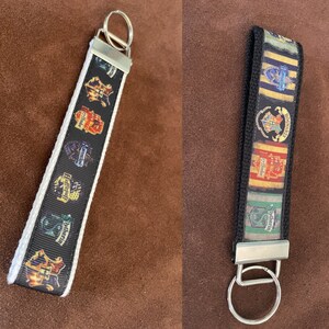 May include: Two Harry Potter house keychains. One keychain has a white background with black ribbon and the Hogwarts house crests. The other keychain has a black background with a colorful ribbon with the Hogwarts house crests.