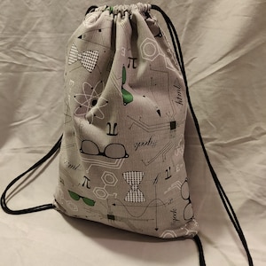 May include: A gray drawstring bag with a science-themed print. The print features glasses, bow ties, atoms, and mathematical equations, including the symbol for pi. The bag has black drawstrings.