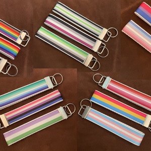 May include: A selection of key fobs displaying pride flag designs. Each fob has a striped pattern in colours representing different identities, including rainbow, bisexual, and transgender. They have silver metal rings.