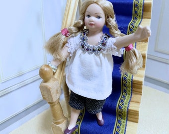 Puede incluir: Una muñeca de porcelana con cabello rubio que lleva una blusa blanca con un collar de cuentas de colores, pantalones negros y cintas para el cabello rojas. La muñeca está de pie en una escalera con un patrón azul y dorado.