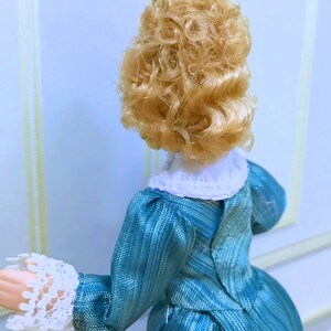 May include: A doll with curly blonde hair wearing a teal blue dress with a textured, striped pattern. The dress features a white lace collar and matching white lace cuffs at the sleeves. The doll is viewed from the back, showing the bodice detail.