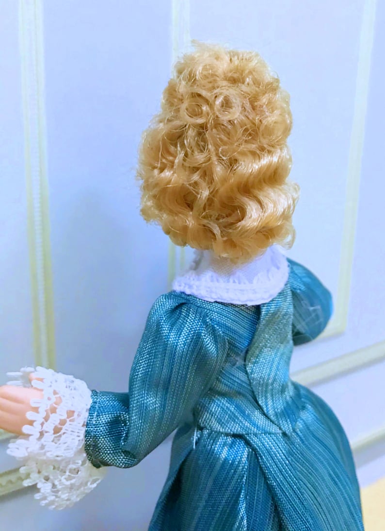 May include: A doll with curly blonde hair wearing a teal blue dress with a textured, striped pattern. The dress features a white lace collar and matching white lace cuffs at the sleeves. The doll is viewed from the back, showing the bodice detail.