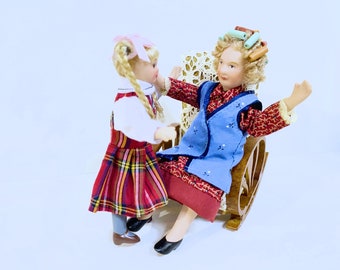 Puede incluir: Dos muñecas de porcelana, una sentada en una mecedora y la otra de pie. La muñeca sentada tiene el pelo en rulos y lleva un chaleco azul sobre un vestido estampado en rojo y blanco. La muñeca de pie lleva un vestido de cuadros rojos y negros con una blusa blanca.