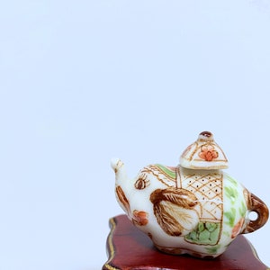 May include: A small, decorative ceramic teapot shaped like an elephant. The teapot is white with brown, green, and orange floral and geometric patterns. It has a small lid and sits on a dark red wooden stand.