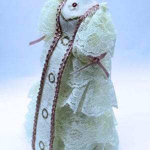 May include: A miniature dress on a mannequin form, crafted from cream-colored lace with tiered ruffles. The garment features a central panel trimmed with pink braided cord, three small gold-toned circular accents, and a single red bead at the neckline.