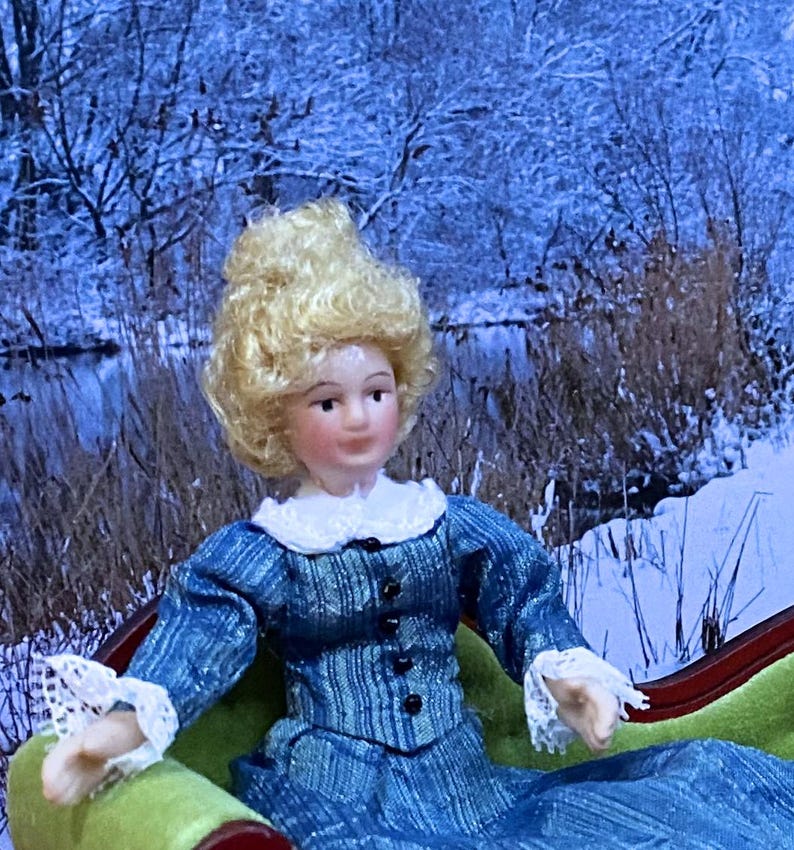 May include: A doll with blonde curly hair, a pale complexion, and a blue long-sleeved dress featuring a white lace collar and white lace cuffs. The dress has a vertical striped pattern and a row of small black buttons down the front bodice.