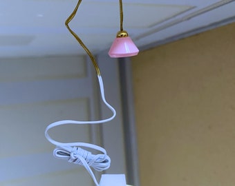 Op de afbeelding: Een wit elektrisch snoer met een witte stekker en een roze lampenkap die hangt aan een gouden metalen armatuur.