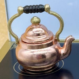 Może przedstawiać: Miniaturowy miedziany czajniczek z czarnym uchwytem i złotymi akcentami. Czajniczek ma zaokrąglony korpus, zakrzywioną wylewkę i pokrywkę z małym pokrętłem. Uchwyt jest łukowaty, a czajniczek stoi na ciemnej powierzchni.
