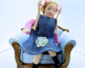 Puede incluir: Una muñeca en miniatura con cabello rubio en trenzas con cintas rosas, vistiendo un vestido de mezclilla azul con un parche floral sobre una camisa de manga larga a rayas rosas y blancas y botas negras, sentada en un pequeño sillón de madera con tela azul.