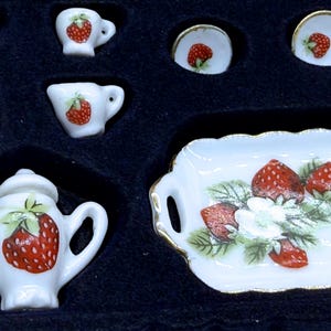 May include: A miniature porcelain tea set featuring a white base with red strawberry patterns and green leaves. The set includes a teapot with a lid, a sugar bowl with a lid, a creamer, two small cups, two saucers, and a rectangular serving tray.