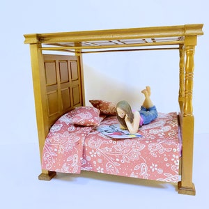 May include: Miniature four-poster bed with a red paisley patterned comforter and pillows. A small figurine is lying on the bed, reading a magazine. The bed frame is made of wood and has a light brown finish.