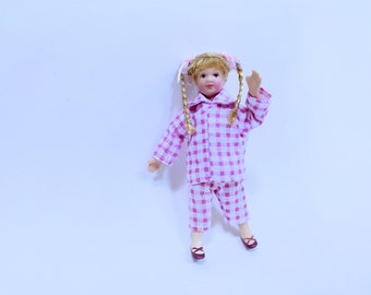 Puede incluir: Una muñeca de porcelana que lleva un pijama a cuadros rosa y blanco con trenzas y una diadema rosa. La muñeca está de pie sobre un fondo blanco.