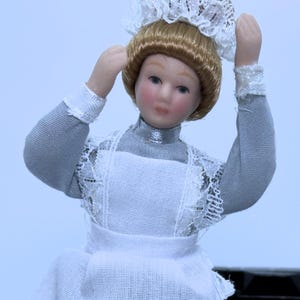 Könnte beinhalten: Eine Porzellanpuppe, die ein weißes Kleid mit einer Spitzenborte und eine weiße Spitzenhaube trägt. Die Puppe hat blonde Haare und blaue Augen.