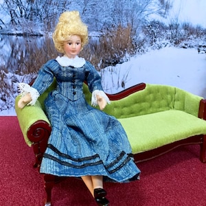 May include: A porcelain doll with blonde hair wearing a blue long-sleeved dress with a white lace collar and cuffs, seated on a miniature green velvet sofa with a dark wood frame. The dress features black buttons and two black horizontal stripes.