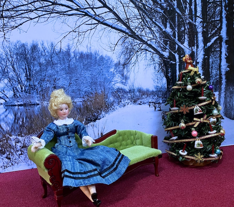May include: A porcelain doll with blonde hair wearing a blue dress with black trim sits on a lime green miniature chaise lounge. Beside the lounge is a decorated Christmas tree with ornaments, ribbons, and a small angel topper.