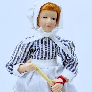 Könnte beinhalten: Eine Porzellanpuppe, die ein weißes Häubchen und ein blau-weiß gestreiftes Kleid trägt. Die Puppe hält einen Holzstab in ihren Händen.