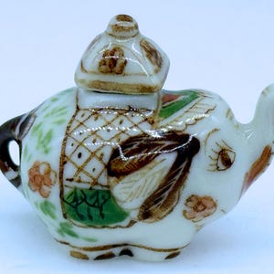 May include: A miniature, white ceramic teapot in the shape of an elephant, complete with a lid. It features brown, green, and orange floral and geometric designs. The handle and spout are brown.