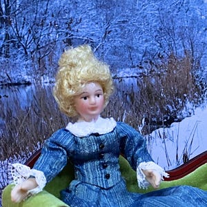 May include: A doll with blonde curly hair, a pale complexion, and a blue long-sleeved dress featuring a white lace collar and white lace cuffs. The dress has a vertical striped pattern and a row of small black buttons down the front bodice.