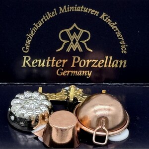 May include: A collection of miniature porcelain kitchenware, including a silver-colored mold, a copper-colored pot, and a copper-colored lidded dish. The items are displayed on a dark blue surface, with the Reutter Porzellan Germany logo visible.