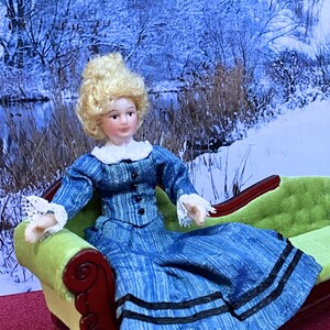 May include: A porcelain doll with blonde hair wearing a blue long-sleeved dress with a white collar, white lace cuffs, and black buttons, seated on a green velvet chaise lounge with a dark wood frame and carved scroll details on the armrest.