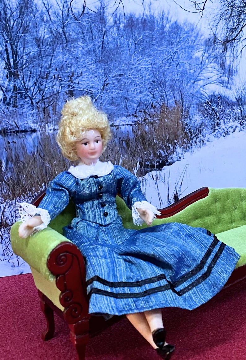 May include: A porcelain doll with blonde hair wearing a blue long-sleeved dress with a white collar, white lace cuffs, and black buttons, seated on a green velvet chaise lounge with a dark wood frame and carved scroll details on the armrest.