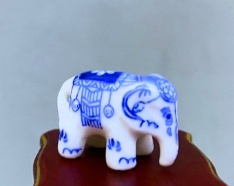 May include: A small, white ceramic elephant figurine with intricate blue patterns. The elephant has a detailed design on its back and head, with a small hole for a string. The figurine is standing on a dark red surface.