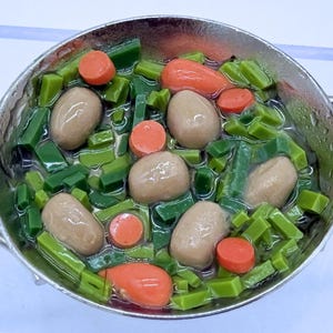 Puede incluir: Una olla en miniatura llena de un guiso con verduras verdes, rodajas de zanahoria naranja y alimentos de forma ovalada de color marrón claro. La olla tiene un asa y un borde plateados. El guiso está sobre un fondo azul claro.